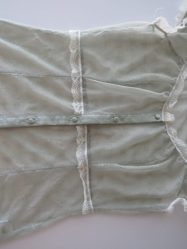 May include: A light sage green sheer mesh blouse featuring a delicate white lace trim along the vertical seams. The garment includes a central button placket with matching sage green fabric-covered buttons and a soft, semi-transparent texture.