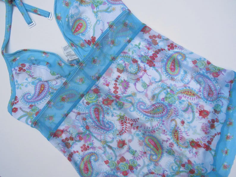 Pu&ograve; includere: Un costume da bagno azzurro in stile allacciato al collo con base bianca e un colorato motivo cachemire e floreale nei toni del rosa, verde e blu. Il capo presenta un pannello in rete trasparente sul petto e spalline regolabili con dettagli bianchi.