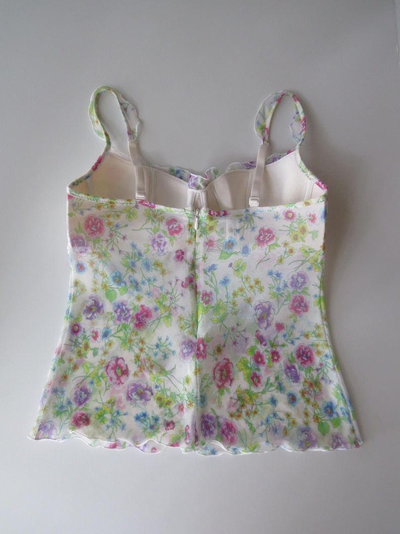 May include: A sleeveless camisole top featuring a delicate floral print with pink, purple, blue, and green flowers on a white background. The garment has thin shoulder straps, a scalloped neckline, and a scalloped hem with a center back zipper.