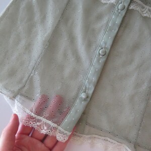 May include: A sage green sheer mesh top featuring a delicate point d'esprit pattern, white lace trim along the edges, and a vertical button placket with matching sage green fabric-covered buttons. The garment has a lightweight, semi-transparent texture.