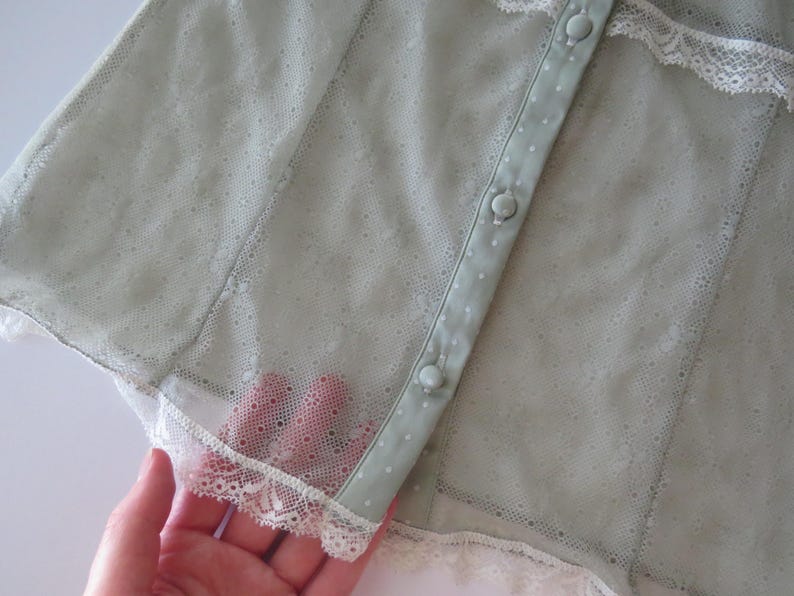 May include: A sage green sheer mesh top featuring a delicate point d'esprit pattern, white lace trim along the edges, and a vertical button placket with matching sage green fabric-covered buttons. The garment has a lightweight, semi-transparent texture.