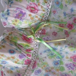 May include: A cream-colored mesh top featuring a vibrant floral print in shades of pink, blue, yellow, and green. The garment includes a vertical strip of white lace trim down the center, accented with two thin, tied ribbons in lime and yellow.