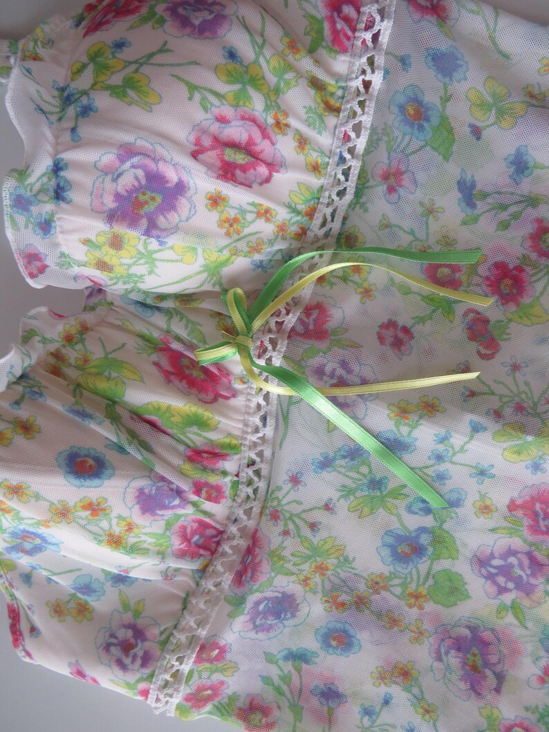 May include: A cream-colored mesh top featuring a vibrant floral print in shades of pink, blue, yellow, and green. The garment includes a vertical strip of white lace trim down the center, accented with two thin, tied ribbons in lime and yellow.