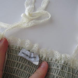 May include: A sage green garment with a smocked bodice and white lace trim along the neckline and shoulder straps. A white fabric tag with the black text "intimissimi" is sewn into the interior neckline of the piece.