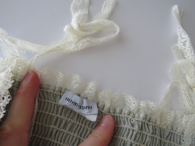 May include: A sage green garment with a smocked bodice and white lace trim along the neckline and shoulder straps. A white fabric tag with the black text "intimissimi" is sewn into the interior neckline of the piece.