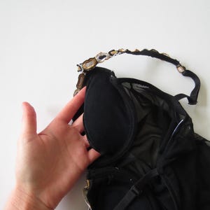 Puede incluir: Lencer&iacute;a negra con cuello halter y sujeci&oacute;n con aros. El sujetador presenta una capa de malla transparente y detalles decorativos dorados y plateados a lo largo del escote y las correas. La prenda est&aacute; sobre un fondo blanco.