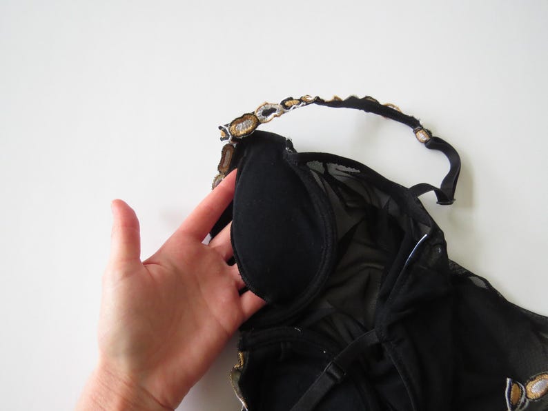 Puede incluir: Lencer&iacute;a negra con cuello halter y sujeci&oacute;n con aros. El sujetador presenta una capa de malla transparente y detalles decorativos dorados y plateados a lo largo del escote y las correas. La prenda est&aacute; sobre un fondo blanco.