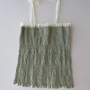 May include: A sage green sleeveless top featuring a smocked, textured bodice with a ruffled hem. The garment includes thin, off-white lace shoulder straps that tie at the top and a matching lace trim along the straight neckline.