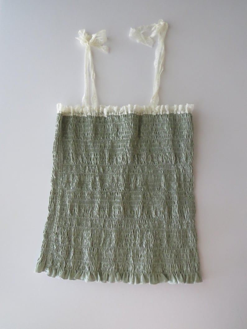 May include: A sage green sleeveless top featuring a smocked, textured bodice with a ruffled hem. The garment includes thin, off-white lace shoulder straps that tie at the top and a matching lace trim along the straight neckline.