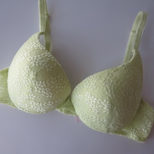 May include: A light lime green bra featuring molded cups with a white floral lace overlay. The garment includes adjustable shoulder straps and a central gore with a small decorative detail, showcasing a structured design with a consistent color palette.