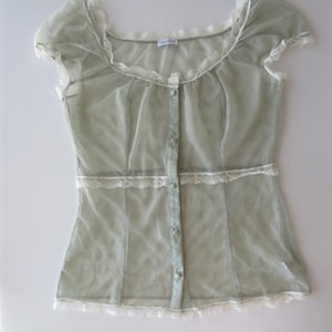 May include: A sage green sheer mesh blouse featuring short cap sleeves, a scoop neckline, and a front button placket. The garment is accented with delicate white lace trim along the neckline, waist, and hem, and includes small matching buttons.