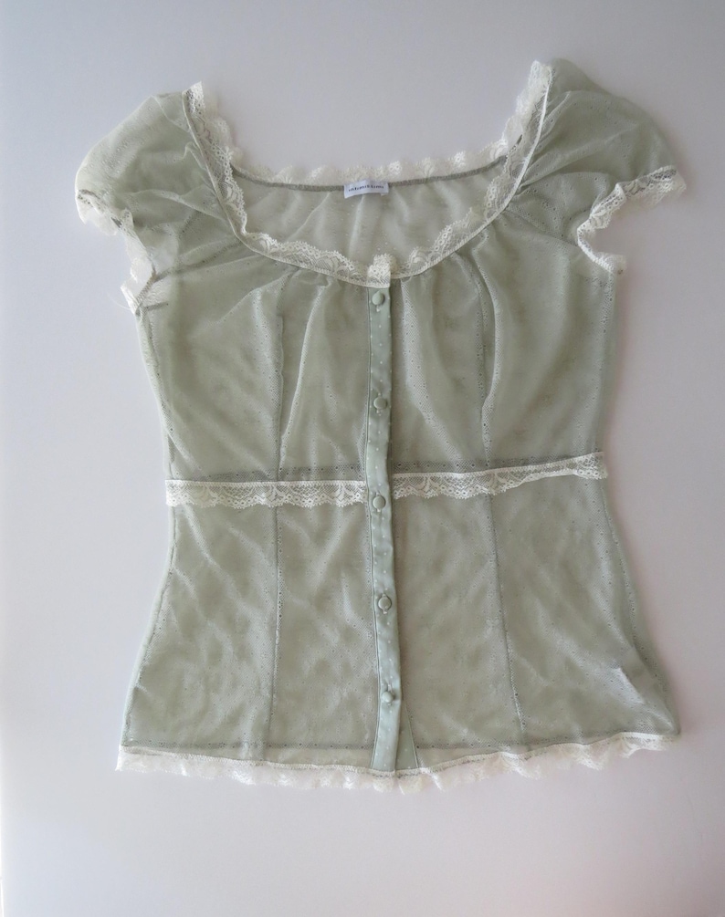 May include: A sage green sheer mesh blouse featuring short cap sleeves, a scoop neckline, and a front button placket. The garment is accented with delicate white lace trim along the neckline, waist, and hem, and includes small matching buttons.