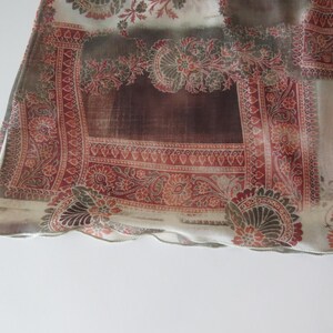 May include: A sheer fabric scarf featuring a repeating pattern of intricate red and brown paisley motifs on a cream background. The rectangular textile has a finished hem along the bottom edge and displays a semi-transparent, lightweight texture.