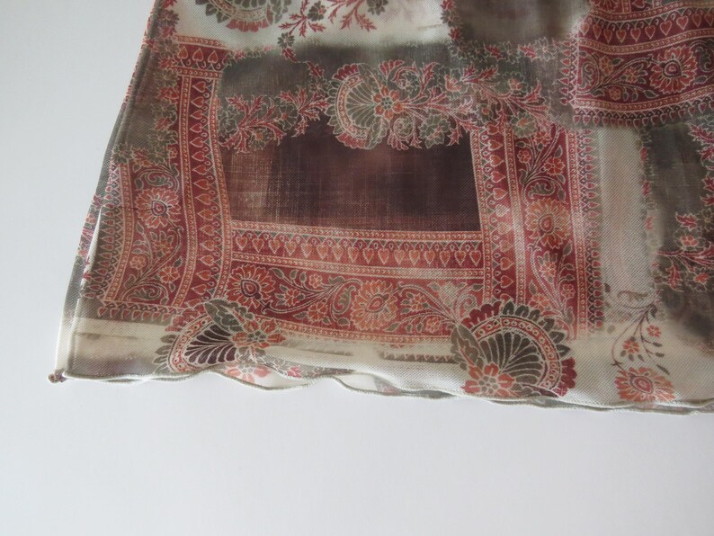 May include: A sheer fabric scarf featuring a repeating pattern of intricate red and brown paisley motifs on a cream background. The rectangular textile has a finished hem along the bottom edge and displays a semi-transparent, lightweight texture.