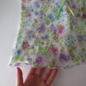 May include: A white sleeveless top featuring a colorful floral print with pink, purple, yellow, and blue flowers and green foliage. The garment has a lettuce-edge hem and a light green ribbon tie detail at the neckline, crafted from a sheer fabric.