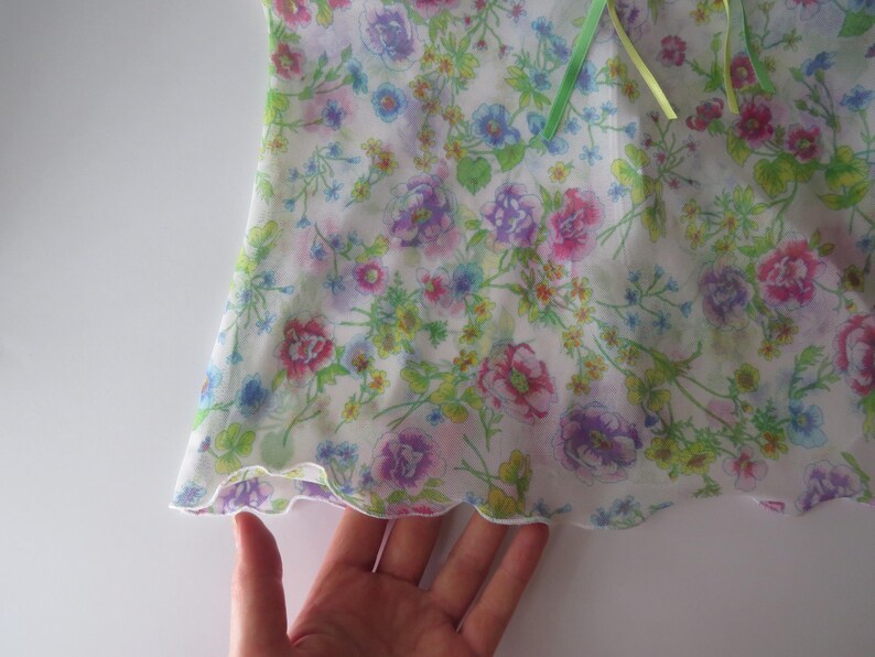 May include: A white sleeveless top featuring a colorful floral print with pink, purple, yellow, and blue flowers and green foliage. The garment has a lettuce-edge hem and a light green ribbon tie detail at the neckline, crafted from a sheer fabric.