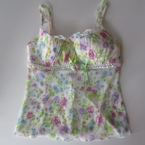 May include: A sleeveless camisole top featuring a white base with a colorful floral print in shades of pink, blue, and green. The garment includes ruffled shoulder straps, a lace trim detail across the chest, and a small lime green ribbon bow.