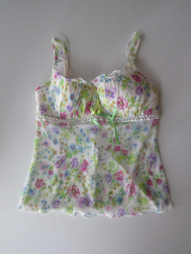 May include: A sleeveless camisole top featuring a white base with a colorful floral print in shades of pink, blue, and green. The garment includes ruffled shoulder straps, a lace trim detail across the chest, and a small lime green ribbon bow.