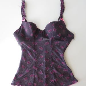 May include: A dark purple bustier featuring a repeating small pink floral pattern, molded cups, and ruffled shoulder straps. The garment includes small pink bows at the base of each strap and a scalloped pink trim along the bottom hemline.