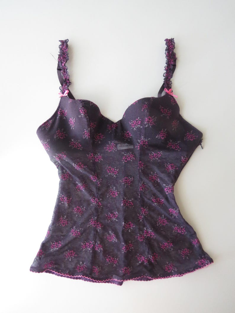May include: A dark purple bustier featuring a repeating small pink floral pattern, molded cups, and ruffled shoulder straps. The garment includes small pink bows at the base of each strap and a scalloped pink trim along the bottom hemline.