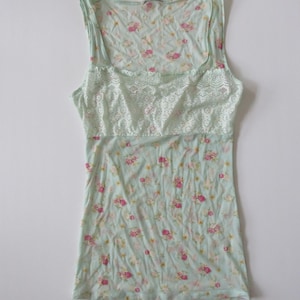 Puede incluir: Camiseta sin mangas azul claro con un estampado floral de flores rosas y amarillas. Tiene un escote cuadrado y un panel de encaje en el pecho. La tela parece suave y ligera.