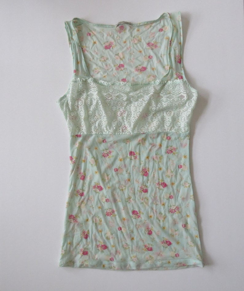 Puede incluir: Camiseta sin mangas azul claro con un estampado floral de flores rosas y amarillas. Tiene un escote cuadrado y un panel de encaje en el pecho. La tela parece suave y ligera.