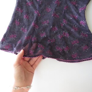 May include: A sheer black mesh top featuring a repeating dark pink floral pattern. The garment is constructed with vertical seams and finished with a delicate, scalloped pink lettuce-edge hem along the bottom border of the fabric.