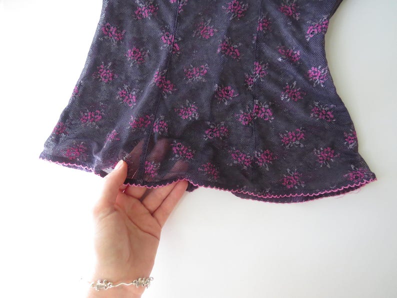 May include: A sheer black mesh top featuring a repeating dark pink floral pattern. The garment is constructed with vertical seams and finished with a delicate, scalloped pink lettuce-edge hem along the bottom border of the fabric.