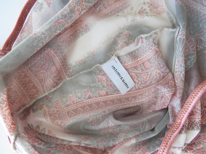 May include: A sheer mesh garment featuring a muted pink, cream, and sage green paisley pattern. The fabric has a fine, semi-transparent texture with a visible white interior tag printed with the brand name intimissimi in black lowercase letters.