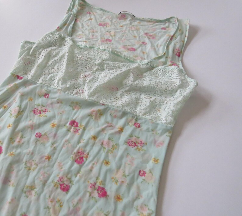 Puede incluir: Camiseta sin mangas con estampado floral azul claro y un panel de encaje blanco en el pecho. La camiseta sin mangas presenta un escote redondo y un estampado floral en rosa y amarillo.