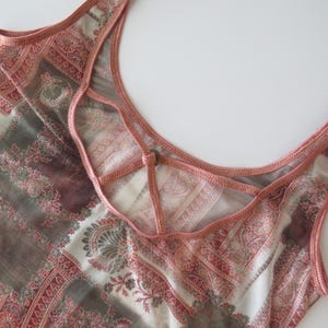 May include: A sleeveless top featuring a multi-colored pattern of muted red, brown, and cream tones. The neckline is accented with a decorative crisscross strap detail in a solid dusty rose fabric that matches the trim along the armholes.