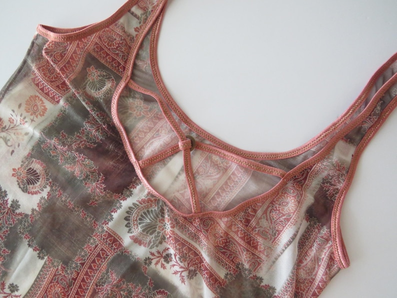 May include: A sleeveless top featuring a multi-colored pattern of muted red, brown, and cream tones. The neckline is accented with a decorative crisscross strap detail in a solid dusty rose fabric that matches the trim along the armholes.