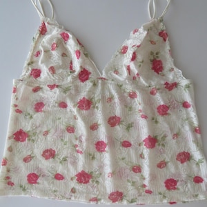 Pu&ograve; includere: Un top a canotta senza maniche realizzato in pizzo bianco con un motivo ripetuto di piccole rose rosa e foglie verdi. Il capo presenta un profondo scollo a V e sottili spalline delicate, mettendo in risalto una trama floreale semi-trasparente.