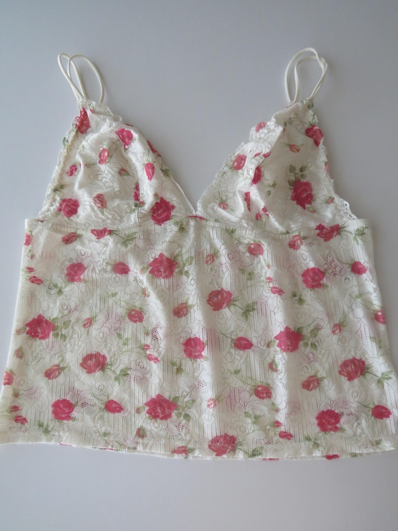 Pu&ograve; includere: Un top a canotta senza maniche realizzato in pizzo bianco con un motivo ripetuto di piccole rose rosa e foglie verdi. Il capo presenta un profondo scollo a V e sottili spalline delicate, mettendo in risalto una trama floreale semi-trasparente.