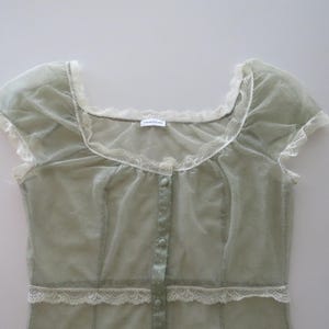 May include: A sage green sheer mesh top featuring short cap sleeves, a square neckline, and a front button placket. The garment is accented with white lace trim along the neckline, sleeves, and waist, and includes a small white brand label.