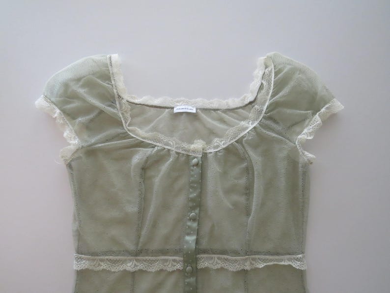 May include: A sage green sheer mesh top featuring short cap sleeves, a square neckline, and a front button placket. The garment is accented with white lace trim along the neckline, sleeves, and waist, and includes a small white brand label.