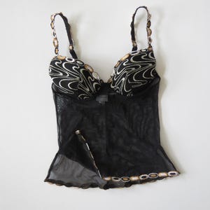 Puede incluir: Top de lencer&iacute;a negro con cuerpo de malla transparente y ribetes decorativos. El top presenta un dise&ntilde;o estampado en blanco y negro en las copas y tirantes, con detalles dorados. Los tirantes son ajustables.