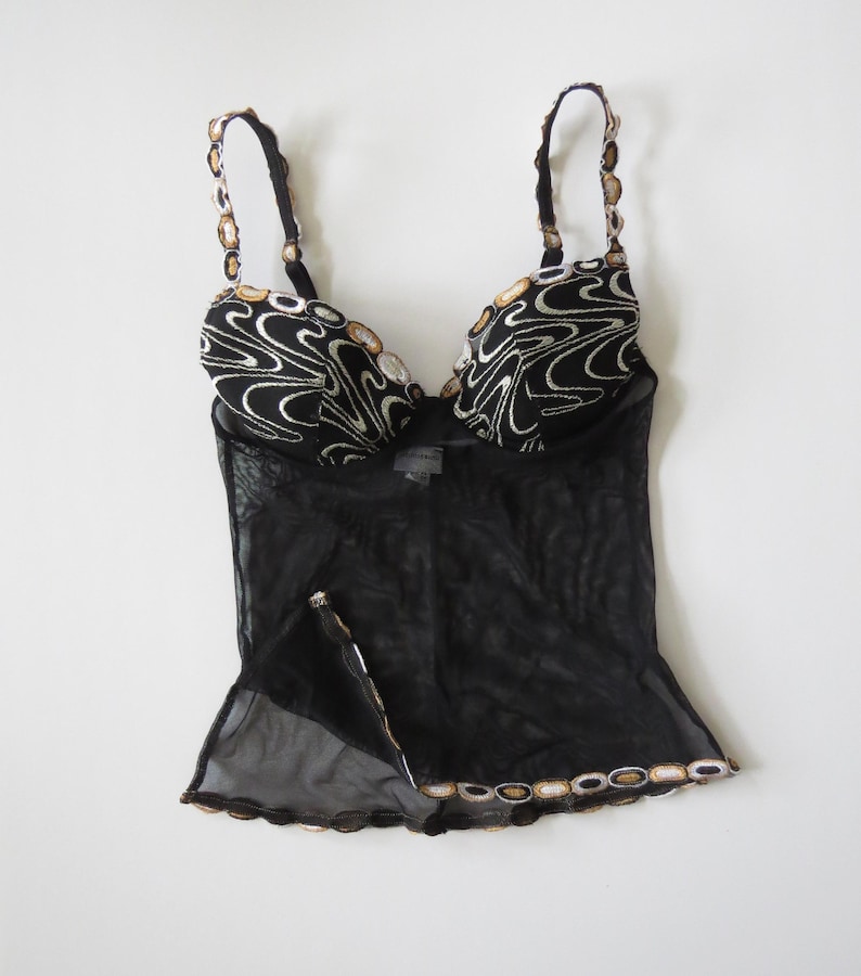 Puede incluir: Top de lencer&iacute;a negro con cuerpo de malla transparente y ribetes decorativos. El top presenta un dise&ntilde;o estampado en blanco y negro en las copas y tirantes, con detalles dorados. Los tirantes son ajustables.
