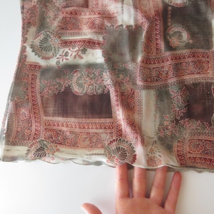 May include: A sheer top featuring a patchwork-style print with deep red, brown, and cream tones. The fabric displays intricate floral and paisley motifs arranged in rectangular sections, finished with a delicate, wavy lettuce-edge hemline.