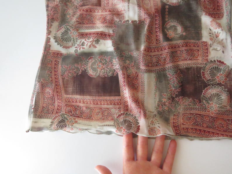 May include: A sheer top featuring a patchwork-style print with deep red, brown, and cream tones. The fabric displays intricate floral and paisley motifs arranged in rectangular sections, finished with a delicate, wavy lettuce-edge hemline.