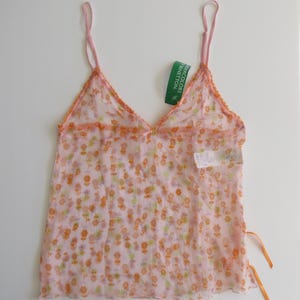 May include: A sheer pink camisole with a floral pattern in orange and green. The top has thin pink straps and orange trim along the neckline. A green tag with white text is attached. The camisole has orange ribbons on the sides.