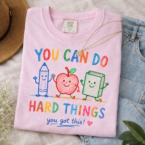 May include: A light pink t-shirt featuring a colorful graphic of a smiling pencil, apple, and book. The text reads "YOU CAN DO HARD THINGS" and "you got this!" with a small heart. The shirt has a Comfort Colors brand label at the inner neckline.