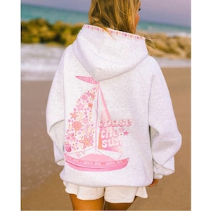 May include: A light gray hoodie with a pink sailboat graphic on the back, featuring the text "chase the sun." The sailboat design includes seashells and starfish. The hood has a pink trim with star patterns.