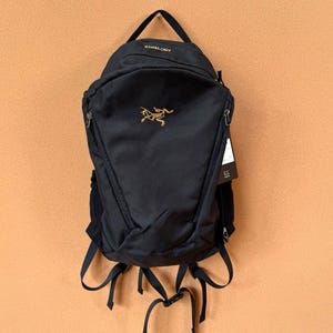 Puede incluir: Mochila negra con asa superior y correas ajustables. La mochila presenta un logotipo dorado y el nombre de la marca "Arc'teryx" en la parte superior. Tiene múltiples compartimentos con cremallera y una etiqueta adjunta.