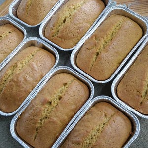 May include: Seven golden-brown loaves of bread are baked in individual rectangular aluminum foil pans. Each loaf features a distinct crack along the center of the top surface, indicating the rise of the dough during the baking process.