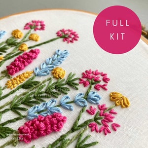 May include: A full embroidery kit featuring a floral design with pink, blue, yellow, and green flowers. The kit includes all the materials needed to complete the project. The text "FULL KIT" is displayed in a pink circle.