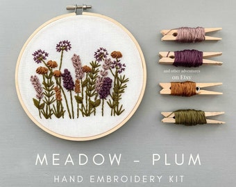 May include: A hand embroidery kit featuring a design of a meadow with purple, brown, and green flowers. The kit includes floss in various colors and a hoop. The text "MEADOW - PLUM" and "HAND EMBROIDERY KIT" is displayed below the hoop.