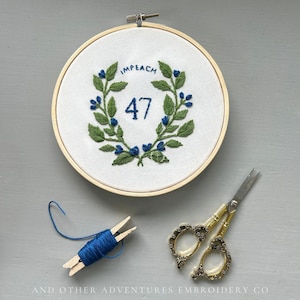 May include: Embroidered hoop art with the word "IMPEACH" above the number "47" surrounded by a green and blue floral wreath. Includes blue thread on a wooden bobbin and gold-handled scissors.