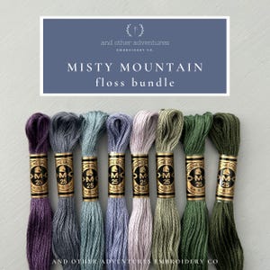 May include: A set of eight DMC embroidery floss spools in shades of purple, blue, green, and gray. The spools are labeled "DMC 25" and "Mouline Special". The text "Misty Mountain floss bundle" is displayed above the spools.