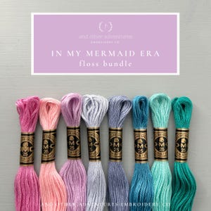 May include: A set of eight embroidery floss spools in shades of pink, lavender, blue, and teal. The spools are labeled "DMC" and "25". The text "In My Mermaid Era floss bundle" is displayed above the spools.